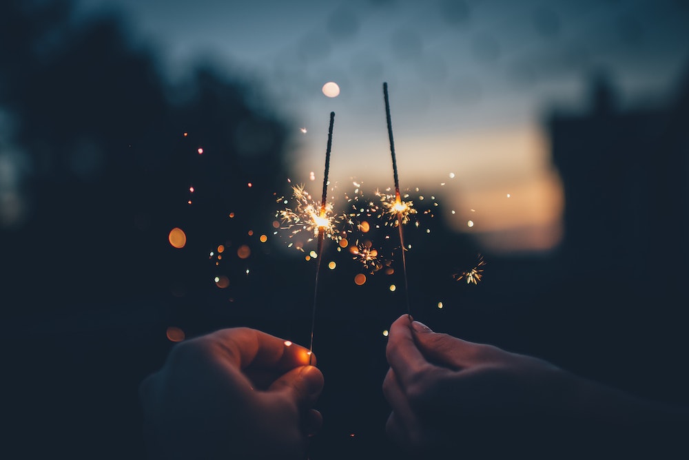 A shallow focus image of a person holding a firecracker as the sun sets in the distance. Photo by Ian Schneider on Unsplash.